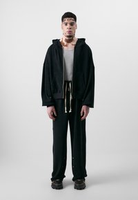 Schwarze Kapuzenjacke mit Reißverschluss und frontalen Taschen, kombiniert mit schwarzen Hosen, die einen elastischen Bund und Kordeln haben. Beide bestehen aus weichem Stoff.