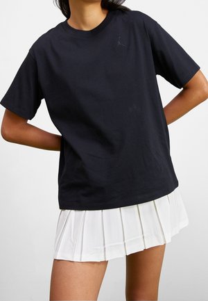 Schwarzes Baumwoll-T-Shirt mit Rundhalsausschnitt und kurzen Ärmeln, mit einem dezenten schwarzen Logo auf der Brust. Kombiniert mit einem weißen Plisseerock.