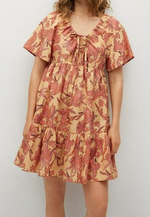 Kobieta w beżowej sukience z krótkim rękawem, ozdobionej czerwonym kwiatowym wzorem paisley, z wiązaniem przy dekolcie.