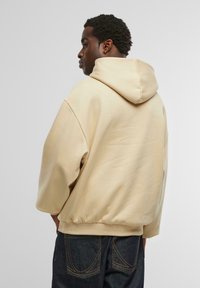 Beige hoodie med en stor huva och lös passform. Har ribbade ärmslut och nederkant. Materialet är slätt med minimal textur. Bärs med mörka jeans.