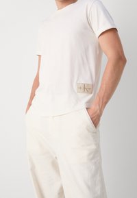 Vit bomullst-shirt med korta ärmar och rund halsringning. Har en brun logotypmärke på v�änster sida. Bärs med ljusa byxor.