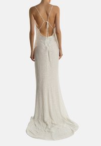 Kvinde iført en gulvlang, hvid, pailletbesat rygfri kjole med tynde stropper, der er bundet i den øverste del af ryggen, stående mod en ensfarvet baggrund.