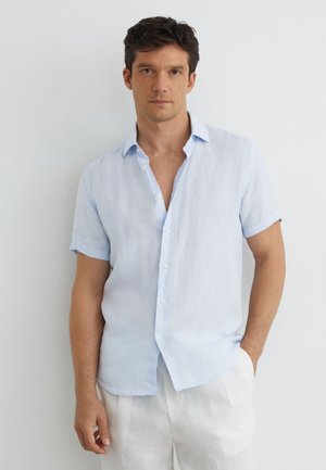 Mann steht an einer weißen Wand, trägt ein hellblaues kurzärmeliges Hemd mit Knöpfen und weiße Hose, eine Hand in der Tasche.