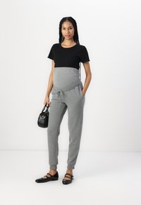 Melnā īsām piedurknēm blūze, pelēkās grūtniecības jogger bikses ar regulējamu jostu un sānu kabatām, apvienotas ar melnām dzīvokļu kurpēm un mazu rokassomu.