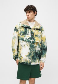 Νεαρός άνδρας που φοράει πολύχρωμο πράσινο, κίτρινο και λευκό ζακέτα με μοτίβο, με γιακά σε ταμπά απόχρωση και σκούρα πράσινα σορτς, κοιτάζει προς τα αριστερά του.