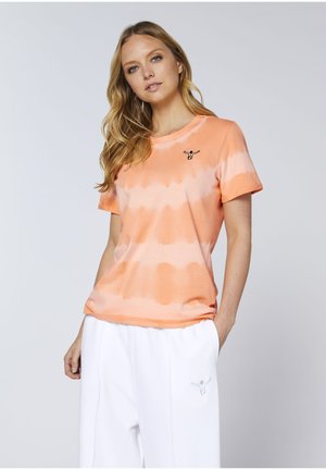 Woman with long blonde hair wearing an orange tie-dye T-shirt and white pants, standing with one hand in pocket against a plain background.