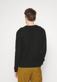 Homme aux cheveux bruns courts portant une chemise noire à manches longues et un pantalon jaune moutarde, debout de dos sur un fond blanc uni.