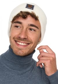 White ribbed beanie with a rectangular logo patch, worn with a blue turtleneck. The background is plain white, highlighting the apparel.