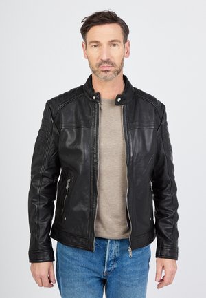 Sort lædermotorcykeljakke med quiltede skulderdetaljer, lynlåslukning foran og to sidelommer, båret over en lys grå skjorte.