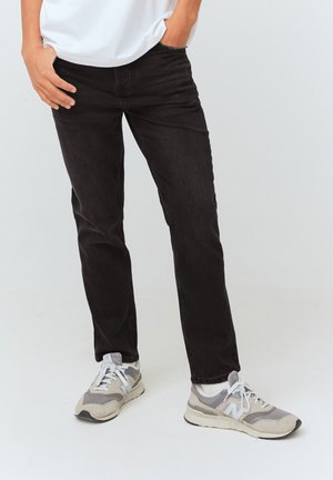 Jeans en denim noir avec une coupe slim, dotés d'un design classique à cinq poches et de coutures discrètes. Portés avec des baskets grises et une chemise blanche.