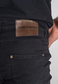 Crne traperice od denima s smešnom kožnom etiketom označenom "GIANNI KAVANAGH." Sadrži metalne kopče i suptilne detalje šivanja.