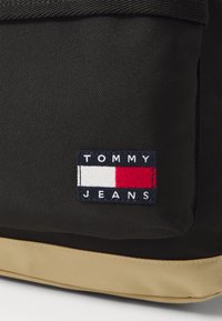 Crni platneni ruksak s pravokutnim aplikacijom s natpisom "TOMMY JEANS", crvenim i bijelim detaljima te smeškastim dnom.