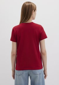Red cotton t-shirt with a simple round neckline and short sleeves, viewed from the back. Paired with light blue denim jeans.