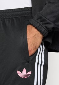 Μαύρο αθλητικό παντελόνι με ελαστική μέση, πλαϊνές τσέπες και λευκές ρίγες. Διαθέτει ροζ κεντημένο λογότυπο στο αριστερό πόδι.