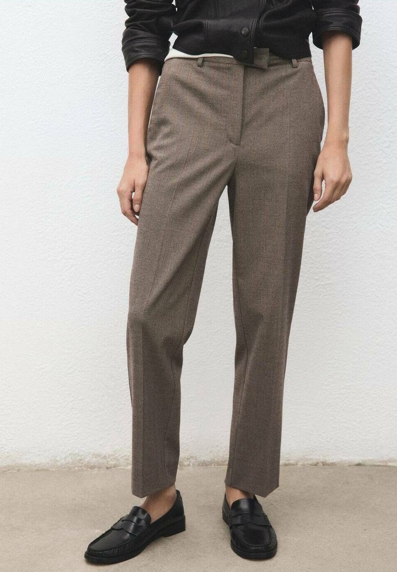 Personne portant un pantalon marron sur mesure, des mocassins noirs et une veste noire, debout contre un mur blanc texturé.