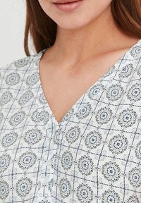Fehér blúz V-nyakkal, geometrikus kék mintával és gombos részletekkel, könnyű anyagból, puha textúrával készítve.