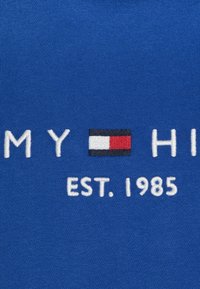 Il tessuto blu presenta il nome del marchio ricamato in bianco e una grafica di bandiera con elementi rossi, bianchi e blu scuro; "EST. 1985" in basso.