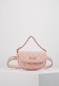 Różowa pikowana skórzana torebka na pasek Guess z złotym łańcuszkowym uchwytem i plakietką z logo, umieszczona na białym piedestale na jednolitym tle.