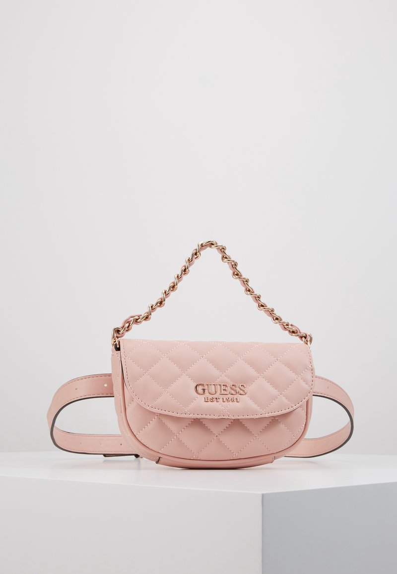 Różowa pikowana skórzana torebka na pasek Guess z złotym łańcuszkowym uchwytem i plakietką z logo, umieszczona na białym piedestale na jednolitym tle.
