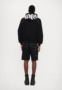 Black hooded sweatshirt with white graphic text on the back, paired with black shorts featuring white stitching and side pockets, worn with black boots.
