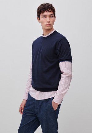 Junger Mann steht mit einer Hand in der Tasche, trägt einen marineblauen Kurzarm-Pullover über einem rosa gestreiften Langarmhemd und blaue Hosen, schaut seitlich.