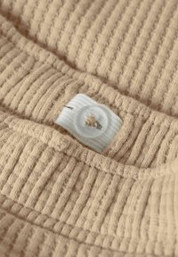 Beige ribbestrikket stoff med en hvit knapp med blomstermotiv. Nærbilde viser tekstur og sømdetaljer.