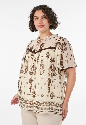 Kvinde med krøllet hår iført en beige bluse med brune blomster- og geometriske mønstre, stående mod en hvid baggrund.