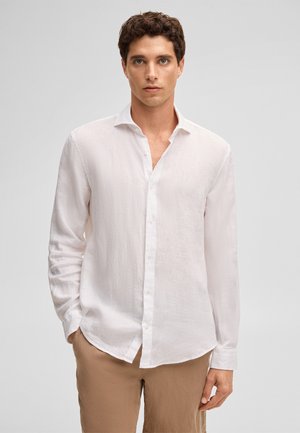 Man with curly dark hair wearing an unbuttoned white long-sleeve shirt and light brown pants, standing against a plain background.