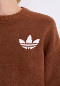 Brun strikket genser med hvit brodert logo med tre striper, rund hals og strukturert stoff.