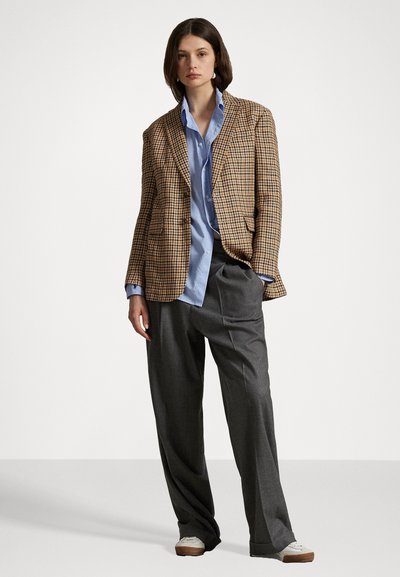 Geruite bruine en gele blazer over een lichtblauw overhemd met knopen. Grijze wijde broek en off-white sneakers maken de outfit compleet.