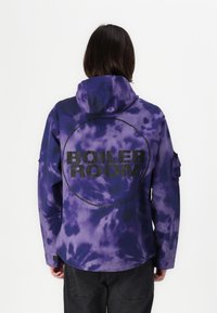 Purple tie-dye jacket with a hood. Features a large black circle design and the text "BOILER ROOM" on the back. Soft, textured fabric.