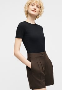 Femme blonde aux cheveux bouclés portant un haut noir à manches courtes et un short marron taille haute à plis, debout devant un fond uni.