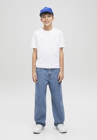 Maglietta bianca a maniche corte, jeans larghi blu chiaro, sneaker bianche e un cappellino blu con logo testuale. Outfit semplice e informale.