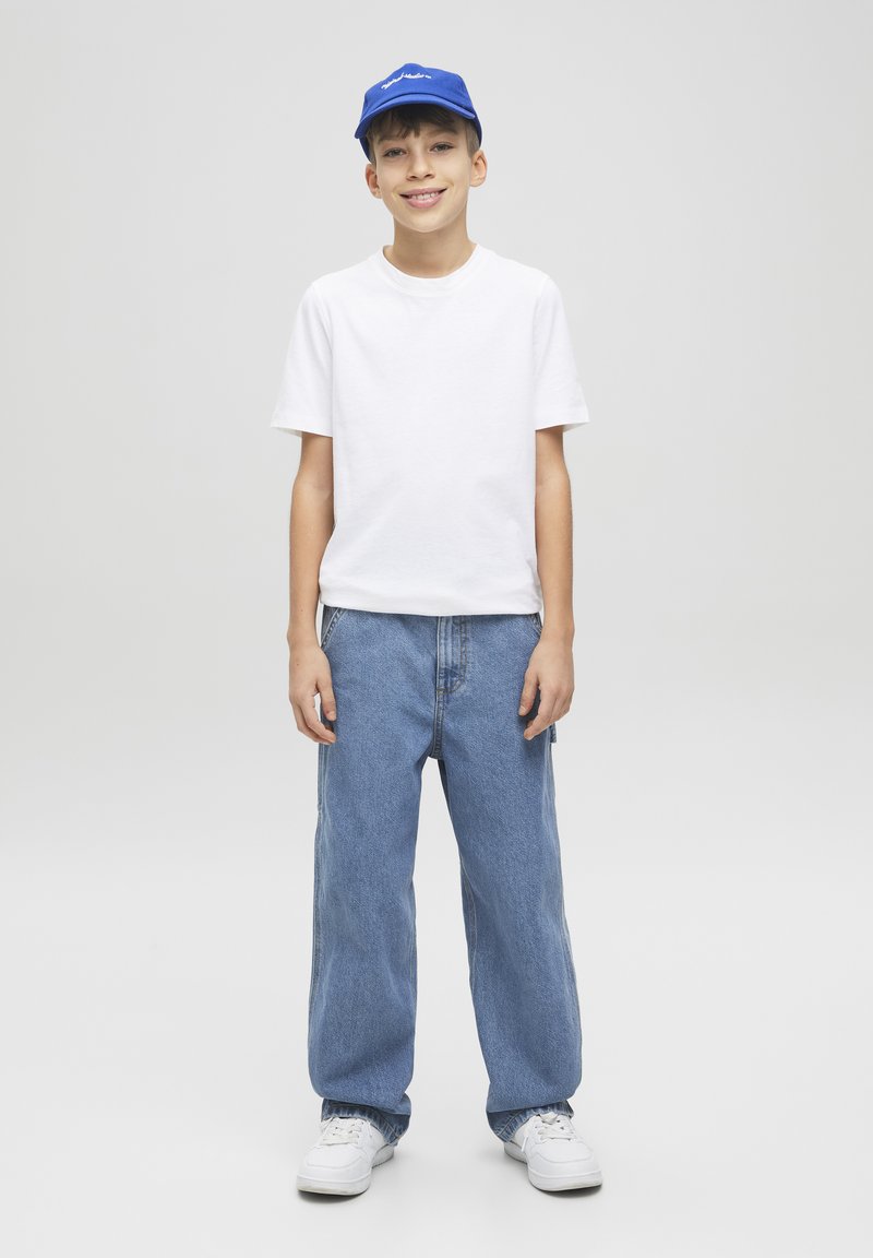 Maglietta bianca a maniche corte, jeans larghi blu chiaro, sneaker bianche e un cappellino blu con logo testuale. Outfit semplice e informale.