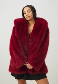 Rød faux fur frakk med stor sjal krage, oversized ermer og en tykk, myk tekstur. Brukes over en svart kjole.