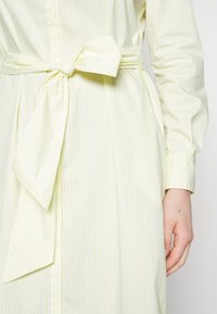 Abito a righe giallo chiaro con maniche lunghe, caratterizzato da una cintura legata in vita. Il tessuto appare liscio e leggero.