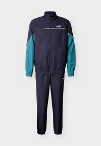 Σκούρο μπλε ναυτικού και τυρκουάζ φόρμα με σακάκι με φερμουάρ, που διαθέτει όρθιο γιακά και ελαστικά μανίκια, συνδυασμένη με στενά παντελόνια, και τα δύο από απαλό ύφασμα.