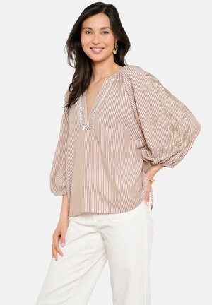 Mujer sonriendo, vestida con una blusa de rayas beige y blancas con bordado floral en las mangas abullonadas y pantalones blancos, de pie con la mano en la cadera.
