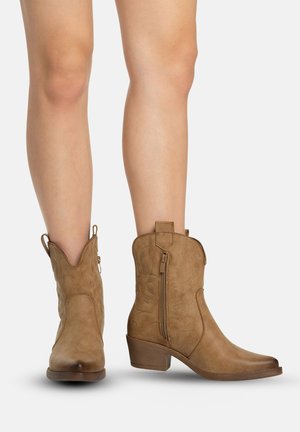 Jambes portant des bottes marron clair en daim style western montant jusqu'au milieu du mollet, avec fermetures éclair latérales, talons blocs bas et coutures décoratives sur fond blanc.