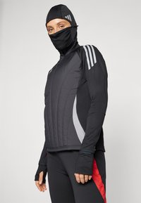 Veste de sport noire avec un devant matelassé, des accents gris et une capuche. Elle présente des ouvertures pour les pouces dans les manches et un design ajusté.