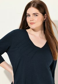 Mörk marinblå V-ringad topp med trekvartslånga ärmar, med subtila plisseringar vid halsringningen. Lätt, texturerad tyg med en avslappnad passform.