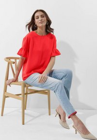 Rote Bluse mit Rüschenärmeln, kombiniert mit hellblauen Jeans. Model sitzt auf einem Holzstuhl und trägt beige Absätze. Minimalistischer Hintergrund.