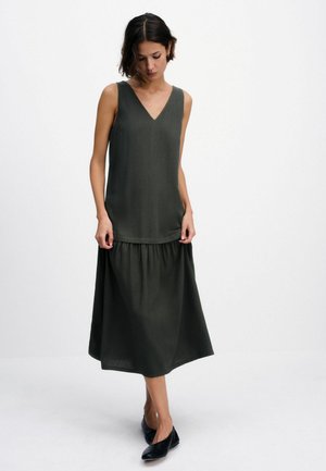 Žena v tmavě zelených šatech bez rukávů s výstřihem do V a nařaseným pasem stojí před bílým pozadím.