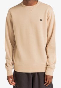 Sweat-shirt beige en tissu doux, avec un col rond, des manches longues, et un petit logo brodé sur la poitrine.