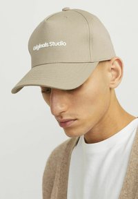Casquette de baseball en tissu beige, arborant le logo brodé "Originals Studio" en blanc. Design arrondi avec une visière légèrement courbée.