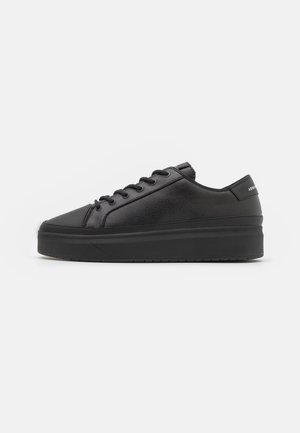 Zapatilla deportiva de cuero negro con textura suave, diseño con cordones y suela gruesa de goma. Presenta un estilo minimalista y puntera suave y redondeada.