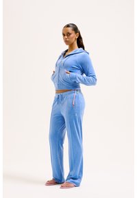 Lichtblauwe fluwelen trainingsoutfit met een aansluitende rits-hoodie en relaxte broek, voorzien van een trekkoord-detail en subtiele merkaccenten.