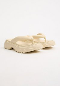 Sandalias de goma en color beige, estilo chancla, con suelas gruesas y texturizadas, presentando un diseño minimalista y una silueta de tiras cruzadas.