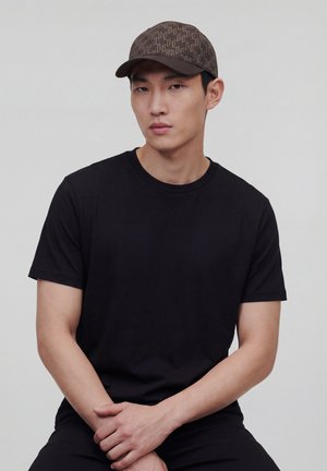 Nuori mies, jolla on kuvioitu ruskea lippalakki ja yksivärinen musta t-paita, istuu kädet yhteen liitettyinä neutraalia harmaata taustaa vasten.