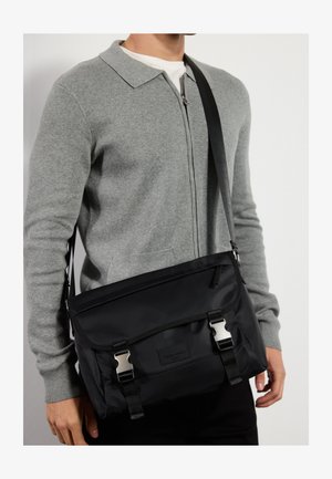 Schwarze Messenger-Tasche aus strapazierfähigem Material, mit silberner Hardware, verstellbarem Tragegurt und doppelten Schnallenverschlüssen an der vorderen Klappe.